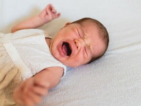 Bebê de vestido branco florido chorando e deitada sobre um lençol branco.