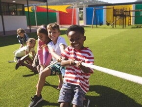 Em um parque ao ar livre, três meninos e duas meninas estão se divertindo em uma brincadeira de cabo de guerra. As crianças, sorridentes, estão puxando a corda.