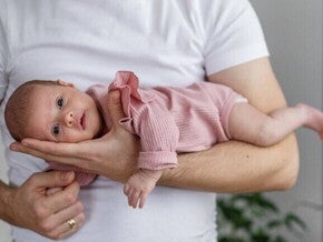 Close-up de um pai segurando um recém-nascido com a barriga para baixo em seu antebraço, em uma posição de "avião".