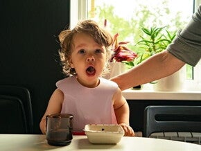 Um bebê com babador sentado à mesa, com a boca aberta, olhando diretamente para a câmera, com a mão de um adulto ao fundo.