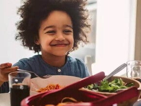 criança sentada a mesa no horário de almoço, sorrindo enquanto segura o talher na mão. Em sua frente estão os pratos com alimentos, incluindo uma salada verde, e há a presença de um adulto do outro lado da mesa.