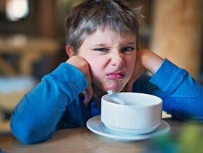 Um menino fazendo uma expressão de desgosto, está apoiando o rosto nas mãos e olhando para a câmera. Na frente dele, em uma mesa de madeira, há uma tigela branca em um pires.