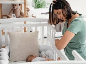 Uma mãe está curvada sobre um berço, olhando para o bebê que dorme. Ela tem a mão na cabeça, transmitindo cansaço, estresse ou preocupação. 