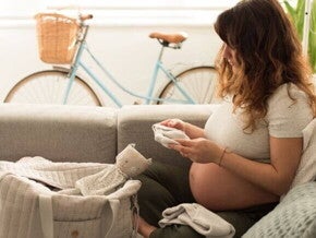 Gestante de cabelo castanho, sentada em um sofá, organizando a bolsa maternidade. Ela segura uma pequena peça de roupa de bebê, com uma bolsa e um boneco de pelúcia ao seu lado. A imagem foca na tarefa de escolher o que levar na bolsa maternidade, enfatizando a importância da preparação.