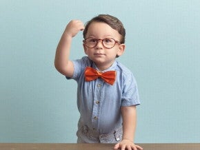 Menino com óculos, gravata vermelha e camisa azul, está apoiado sobre uma mesa, com a mão direita levantada em punho, sob um fundo azul.