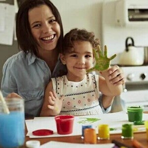 Sentadas à mesa estão uma menina e uma mulher. Espalhadas pela superfície temos tintas de diversas cores e papéis. A mão da criança está toda pintada de verde e as duas estão alegres. A imagem remete a brincadeiras para fazer com crianças mais crescidinhas Sentadas à mesa estão uma menina e uma mulher. Espalhadas pela superfície temos tintas de diversas cores e papéis. A mão da criança está toda pintada de verde e as duas estão alegres. A imagem remete a brincadeiras para fazer com crianças mais crescidinhas