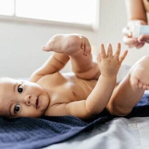 Um bebê deitado de fralda em um trocador sobre uma toalha azul-escura, com as pernas levantadas. Na parte superior direita, mãos desfocadas de um adulto seguram um tubo de creme, preparando-se para aplicar pomada para assadura por alergia alimentar ou de contato.