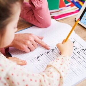 Uma mão adulta aponta para um caderno quadriculado onde uma criança está praticando a escrita das letras "A" e "a" com um lápis. Uma mão adulta aponta para um caderno quadriculado onde uma criança está praticando a escrita das letras "A" e "a" com um lápis.