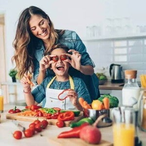 Uma mãe e sua criança estão na cozinha, com diversos alimentos sobre uma bancada. Ela segura dois tomates pequenos na altura dos olhos da criança e ambas estão sorrindo Uma mãe e sua criança estão na cozinha, com diversos alimentos sobre uma bancada. Ela segura dois tomates pequenos na altura dos olhos da criança e ambas estão sorrindo