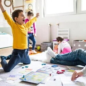 16 atividades lúdicas para fazer em casa com as crianças Sala com um adulto, onde só é possível ver suas pernas usando calça jeans e meia. Na mesma sala, estão três crianças. Uma garota com cerca de 1 ano de idade em segundo plano, branca, de cabelos curtos, de costas brincando com uma boneca. Outra garota com cerca de quatro anos, branca, cabelos loiros presos, calça jeans e blusa de manga comprida. Ela parece estar feliz dançando. A criança em destaque é um menino, branco, com cabelos curtos, de braços abertos comemorando uma vitória em um jogo