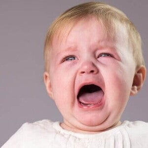 bebê loiro de olhos claros chorando com a boca aberta e expressão de desconforto.