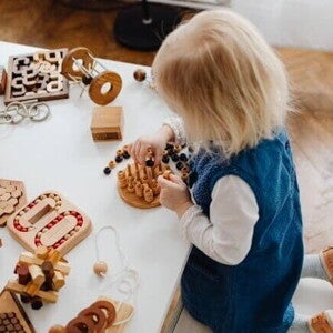 Uma criança está sentada em uma mesa branca e se diverte com uma série de brinquedos sensoriais de madeira. A cena foca no envolvimento da criança com as diferentes texturas e formas, ressaltando o tema de brincadeiras sensoriais. Uma criança está sentada em uma mesa branca e se diverte com uma série de brinquedos sensoriais de madeira. A cena foca no envolvimento da criança com as diferentes texturas e formas, ressaltando o tema de brincadeiras sensoriais.