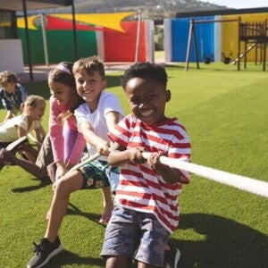 Como passar as brincadeiras do folclore brasileiro para seus filhos? Em um parque ao ar livre, três meninos e duas meninas estão se divertindo em uma brincadeira de cabo de guerra. As crianças, sorridentes, estão puxando a corda.