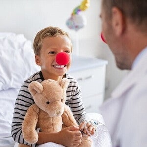 Um menino sorridente com um nariz de palhaço vermelho e segurando um ursinho de pelúcia está sentado em uma cama de hospital, olhando para um médico de costas para o observador. O médico também usa um nariz de palhaço vermelho. Um menino sorridente com um nariz de palhaço vermelho e segurando um ursinho de pelúcia está sentado em uma cama de hospital, olhando para um médico de costas para o observador. O médico também usa um nariz de palhaço vermelho.