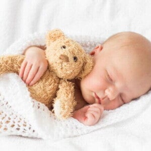Bebê dormindo tranquilamente abraçado a um ursinho de pelúcia, enrolado em uma coberta branca de crochê