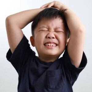 Um close-up mostra o rosto de um menino franzindo a testa, com os olhos fechados e as mãos na cabeça, demonstrando intensa frustração. Seus dentes estão cerrados, e a expressão facial captura um momento de grande descontentamento e dificuldade em lidar com suas emoções. O fundo branco direciona o foco para a reação emocional da criança.