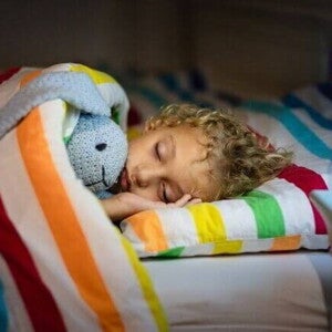 Criança dormindo tranquilamente abraçada a um bicho de pelúcia, coberta por um edredom listrado e colorido.