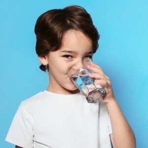Um menino está bebendo água de um copo transparente, olhando para a câmera, sob um fundo azul. Um menino está bebendo água de um copo transparente, olhando para a câmera, sob um fundo azul.