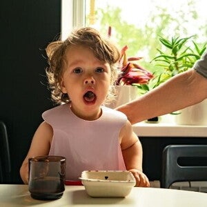 Um bebê com babador sentado à mesa, com a boca aberta, olhando diretamente para a câmera, com a mão de um adulto ao fundo. Um bebê com babador sentado à mesa, com a boca aberta, olhando diretamente para a câmera, com a mão de um adulto ao fundo.