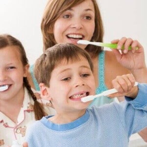 Uma foto de uma mãe e seus filhos, sorrindo e escovando os dentes juntos no banheiro. A imagem sugere uma maneira divertida de como ensinar crianças a escovar os dentes. Uma foto de uma mãe e seus filhos, sorrindo e escovando os dentes juntos no banheiro. A imagem sugere uma maneira divertida de como ensinar crianças a escovar os dentes.