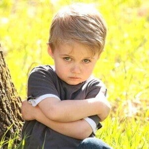 Um menino está encostado em uma árvore, com os braços cruzados e uma expressão de desafio no rosto. A postura e a expressão do menino exemplificam a teimosia e a afirmação de independência características da fase do não infantil.