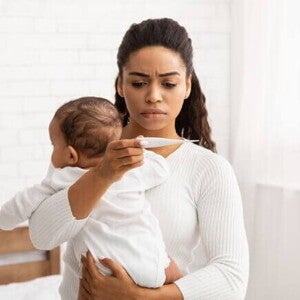 Uma mulher segura um bebê nos braços, com feição preocupada, enquanto verifica a temperatura do bebê com um termômetro. Uma mulher segura um bebê nos braços, com feição preocupada, enquanto verifica a temperatura do bebê com um termômetro.