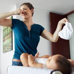 Uma mulher em pé, segurando uma fralda em uma mão e tapando o nariz com a outra mão, com um bebê deitado em sua frente