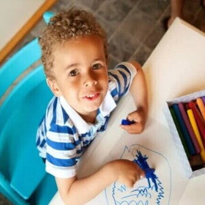 criança sentada em cadeira e mesa infantil com giz de cera azul em mão, desenhando em folha de papel, outras cores de giz disponíveis sobre a mesa criança sentada em cadeira e mesa infantil com giz de cera azul em mão, desenhando em folha de papel, outras cores de giz disponíveis sobre a mesa