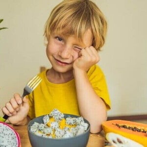Um menino sentado à mesa com um prato de frutas variadas segurando um garfo e piscando um olho. A imagem representa um lanche da tarde para crianças com frutas saudáveis.