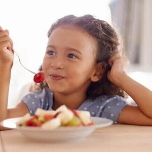 Lanchinho: o que a criança pode comer à noite para dormir bem Representando bons hábitos alimentares, uma criança come seu lanchinho: várias frutas picadas em um prato.