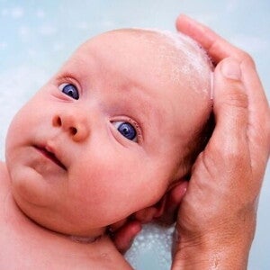 Um close-up de um bebê recém-nascido, deitado na água do banho com espuma. Uma mão adulta com sabão na ponta dos dedos está suavemente lavando a cabeça do bebê. Um close-up de um bebê recém-nascido, deitado na água do banho com espuma. Uma mão adulta com sabão na ponta dos dedos está suavemente lavando a cabeça do bebê.