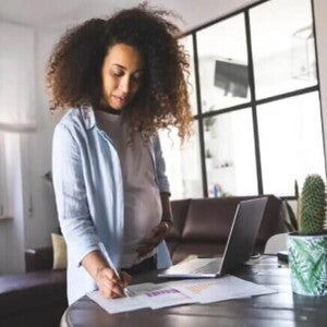 Uma mulher grávida escreve em alguns papéis na mesa da sala de casa, enquanto está de licença-maternidade. Uma mulher grávida escreve em alguns papéis na mesa da sala de casa, enquanto está de licença-maternidade.