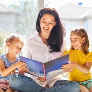 Uma mulher sorridente está sentada lendo um livro para duas meninas pequenas sentadas ao seu lado. Uma mulher sorridente está sentada lendo um livro para duas meninas pequenas sentadas ao seu lado.