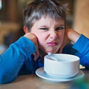 Meu filho não quer comer Um menino fazendo uma expressão de desgosto, está apoiando o rosto nas mãos e olhando para a câmera. Na frente dele, em uma mesa de madeira, há uma tigela branca em um pires.