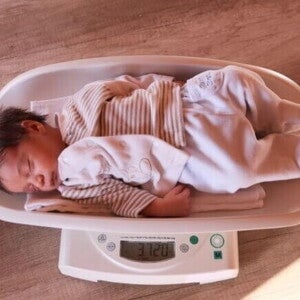 Um bebê recém-nascido dormindo sobre uma balança digital que mostra o peso. A imagem ilustra o monitoramento do peso do bebê, um tema central para a perda de peso neonatal.