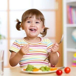 Uma menina pequena e sorridente está sentada à mesa com um prato saudável para crianças na frente dela, que contém vegetais, enquanto ela segura uma colher.