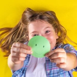 Uma criança deitada e sorrindo, segurando um círculo de papel verde com um rosto sorridente desenhado. Uma criança deitada e sorrindo, segurando um círculo de papel verde com um rosto sorridente desenhado.
