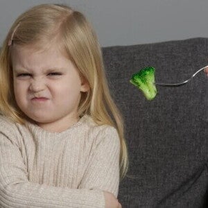 Uma menina pequena está sentada em um sofá cinza com os braços cruzados e uma expressão de desagrado ou nojo no rosto, franzindo as sobrancelhas e a boca. 