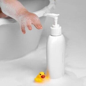 Mão de um bebê se esticando para alcançar um frasco de shampoo ao lado de um pato de borracha.