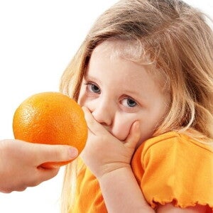 Uma mão oferece uma laranja a uma menina, que cobre a boca com a mão e olha para a câmera com uma expressão de repulsa ou aversão. Uma mão oferece uma laranja a uma menina, que cobre a boca com a mão e olha para a câmera com uma expressão de repulsa ou aversão.