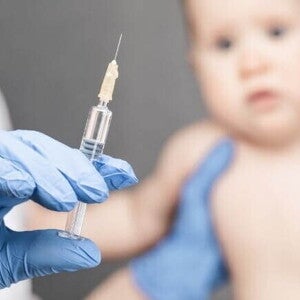 Mão com luva cirúrgica segurando seringa com a vacina da coqueluche, enquanto bebê aguarda a aplicação