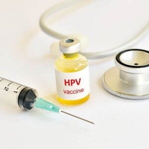 Vista superior de um frasco de vacina rotulado "HPV vaccine" com líquido amarelado, colocado entre uma seringa hipodérmica e um estetoscópio médico, todos dispostos sobre uma superfície branca.