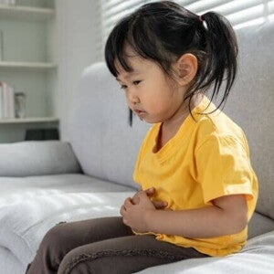 Uma criança sentada no sofá com a mão na barriga, expressando dor. A imagem representa o mal-estar causado por verme infantil.