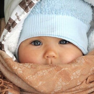 Um close-up de um bebê usando um chapéu azul e um cobertor marrom enrolado em volta do rosto, mostrando como agasalhar um bebê para o frio e como vestir um bebê de acordo com a temperatura. Um close-up de um bebê usando um chapéu azul e um cobertor marrom enrolado em volta do rosto, mostrando como agasalhar um bebê para o frio e como vestir um bebê de acordo com a temperatura.