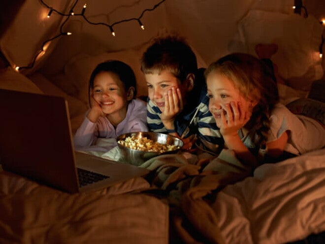 Três crianças, sendo um menino e duas meninas, deitados assistindo no notebook enquanto comem pipoca. Ao fundo, luminárias em uma cabana.