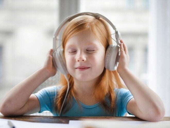 Uma menina de olhos fechados, usando fones de ouvido e sorrindo levemente, com as mãos próximas aos fones.