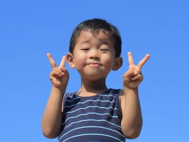 Um menino japonês sorrindo, faz um sinal de "V" com as duas mãos, com o céu azul claro ao fundo.