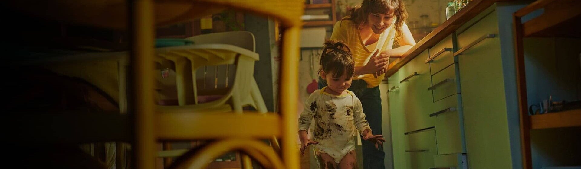 Mãe vestida com uma blusa amarela, encostada na bancada da cozinha, observa, sorridente, a filha que está suja com tinta nas mãos, nas pernas e na roupa 