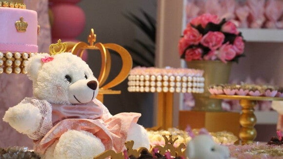 Um urso de pelúcia branco com uma coroa dourada e vestido rosa sentado em uma mesa de sobremesas em uma festa de chá de bebê.