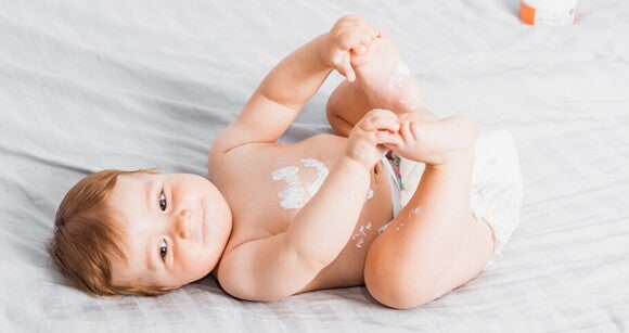 Bebê deitado em uma cama usando fralda e com creme hidratante na barriga